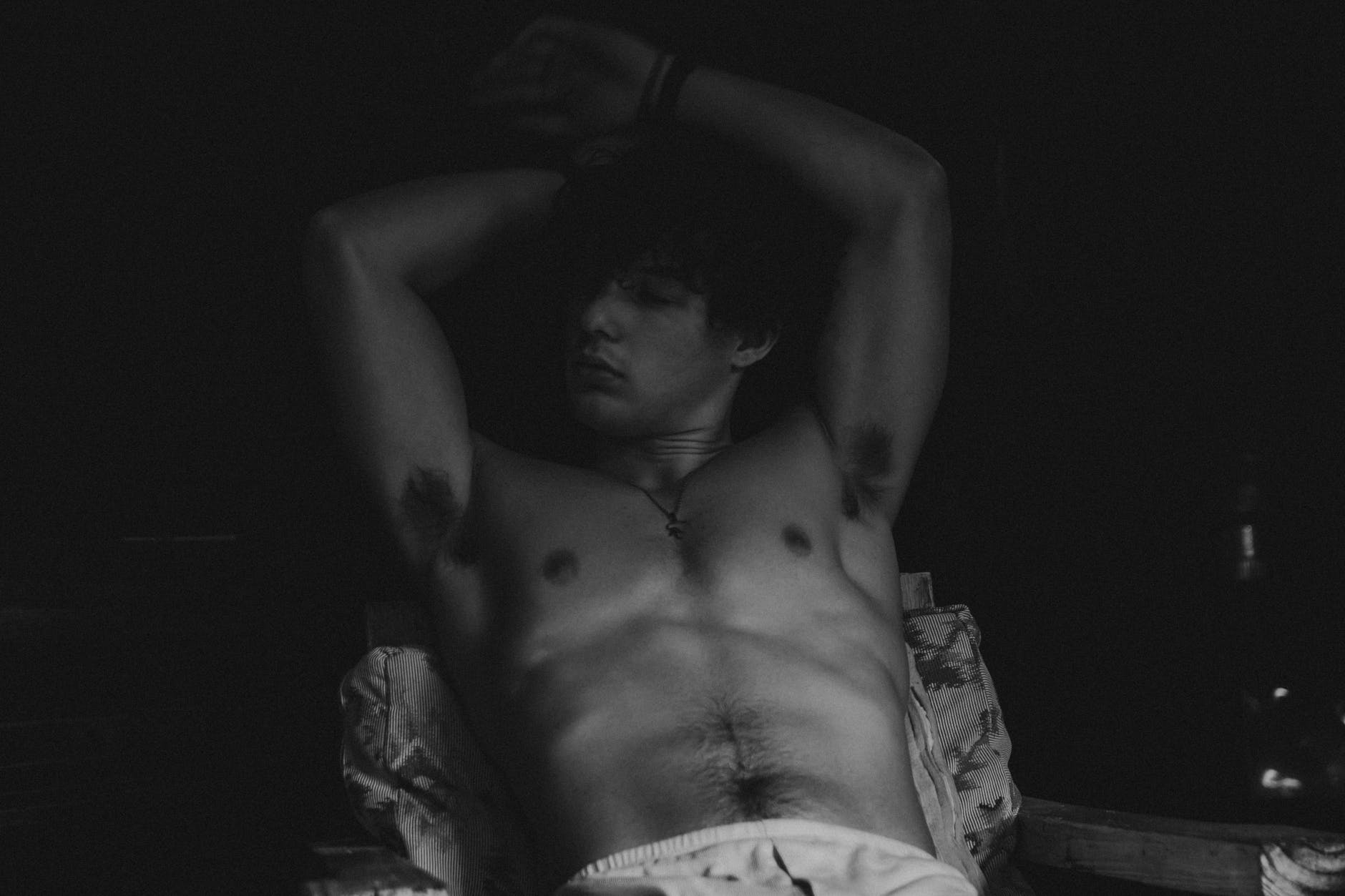 grayscale photo of a shirtless man leaning on a headboard